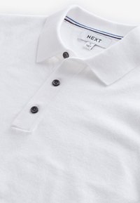 Camiseta polo blanca con un tejido texturizado, tapeta de botones con tres botones negros y un cuello con rayas azules y rojas en la banda interior.