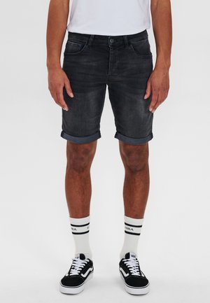 Shorts di jeans in lavaggio scuro con risvolti arrotolati, dotati di tasche frontali e chiusura con bottone. Abbinati a calzini bianchi e sneaker nere.