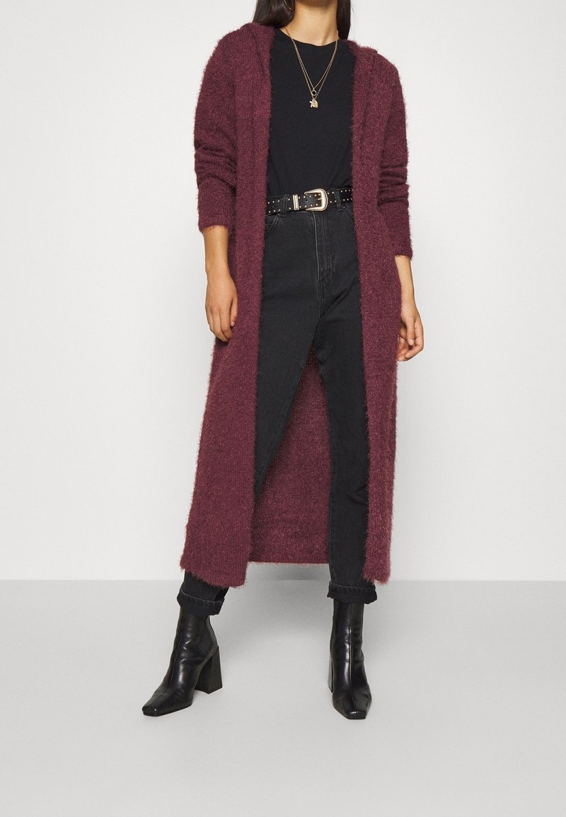 Maglione lungo bordeaux in materiale soffice, con apertura frontale. Indossato sopra una maglia nera e jeans neri, abbinato a stivaletti neri.