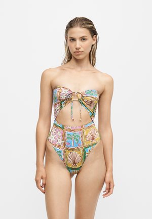 Femme portant un maillot de bain bustier, taille haute, avec une découpe à l'avant et un imprimé tropical coloré de coquillages et d'étoiles de mer sur fond blanc.