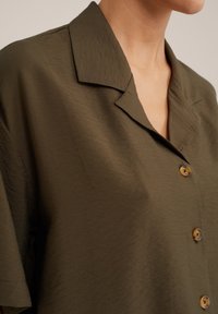 Primo piano di una persona che indossa una camicia verde oliva con bottoni, colletto a punte e maniche corte.