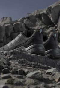 Pair of black athletic shoes with thick soles resting on rocky terrain under clear sky, emphasizing durability for outdoor use.