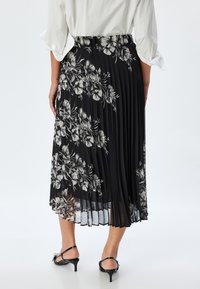 Jupe plissée noire avec un imprimé floral en blanc et gris. Dotée d'une taille haute et d'une texture transparente. Portée avec des sandales à talons noires.