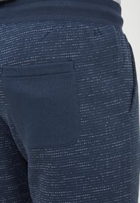 Marinblå joggers i mjukt tyg med ett texturerat mönster med vita prickar och en enda bakficka. Synligt elastiskt midjeband.