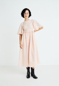 Vestido de encaje rosa claro con mangas cortas abullonadas, cintura ajustada y una falda fluida, combinado con botines negros hasta el tobillo.