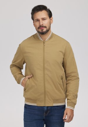 Mężczyzna o ciemnych włosach i brodzie, ubrany w beżową kurtkę typu bomber z mankietami w białe paski oraz niebieskie dżinsy, z lewą ręką w kieszeni kurtki.