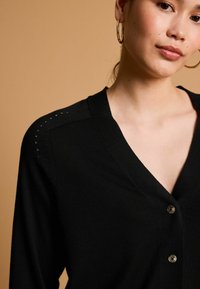 Černý cardigan s V-výstřihem, s knoflíkovým zapínáním a detailními stehy na ramenou, vyrobený z měkké pleteniny.