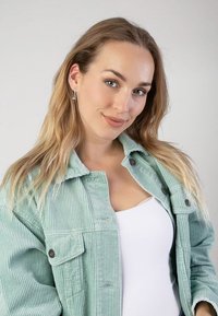 Mintgroene ribfluwelen jas met zichtbare verticale ribbels, knoopsluiting, gedragen over een witte top; model heeft lang, golvend haar en zilveren oorbellen.