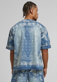 Short-sleeve denim shirt with intricate light and dark blue paisley patterns, frayed hem, and a relaxed fit. Features contrasting white stitching.