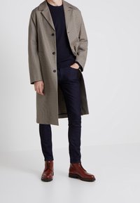 Kontrollerad beige ytterrock med stor krage, svart tröja, mörkblå åtsittande jeans och bruna ankelstövlar med snörning.