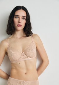 Svetloružová čipkovaná podprsenka s kvetinovými detailami, sieťovými panelmi, nastaviteľnými ramienkami a mašličkou v strede. Hladký povrch a jemná textura.