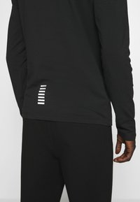 Maglietta nera a maniche lunghe con scollo rotondo, vestibilità slim e dettaglio a strisce riflettenti sulla parte bassa della schiena. Tessuto morbido e liscio.