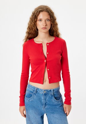Red ribbed cardigan with long sleeves and a cropped design, featuring gold-tone buttons down the front. Paired with light blue jeans.