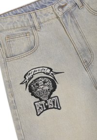 Jasnoniebieskie dżinsy z łatą przedstawiającą ryczącego tygrysa oraz banerem z napisem „Art for Life” nad napisem „EST. 1971” na udzie.