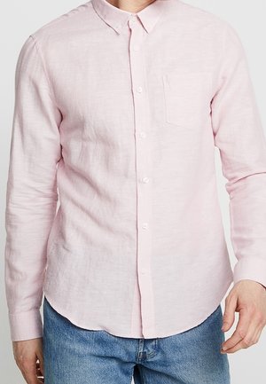 Homme portant une chemise rose pâle à manches longues avec une poche poitrine et un jean bleu, debout devant un fond uni.