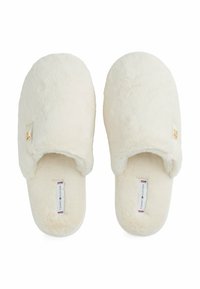 Witte pluche instapslippers met het Tommy Hilfiger-label aan de binnenkant en een klein gouden logo aan de buitenkant van elke slipper.