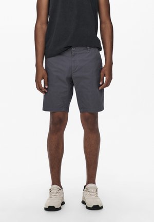 Shorts en coton gris à coupe décontractée, avec une fermeture à boutons, des poches avant et un ourlet cousu. Portés avec un T-shirt noir et des baskets.