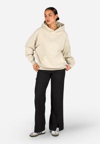 Sweatshirt con cappuccio crema e tasca frontale, vestibilità comoda, abbinato a pantaloni neri con spacchi laterali e sneakers grigio-bianche.