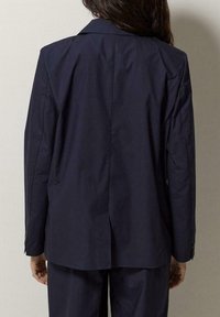 Blazer su misura blu navy con colletto a punta, maniche lunghe e uno spacco singolo sul retro. Tessuto liscio con una leggera lucentezza.