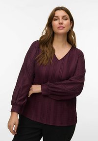 Femme aux longs cheveux bruns portant un chemisier texturé à manches longues violet foncé et un pantalon noir sur un fond blanc uni.