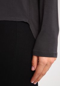 Maglione grigio a maniche lunghe con uno spacco laterale, caratterizzato da una trama morbida. Mano appoggiata su un indumento nero, a mostrare un design stratificato.