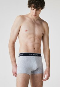 Boxer shorts en coton gris avec une ceinture à logo Lacoste noir, présentant une coupe ajustée et une texture lisse. Le modèle se tient avec une expression neutre.