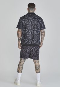Čierna kvetinová košeľa s krátkym rukávom a zodpovedajúce šortky majú tmavý, textúrovaný vzor. Biely tenisky a biele ponožky dotvárajú outfit.