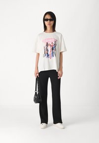 T-shirt graphique blanc avec un design rétro "Clueless", associé à un pantalon noir côtelé et des baskets blanches, tenant un sac noir.
