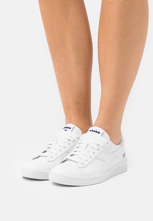 Baskets en cuir blanc avec semelles plates, design à lacets et marquage discret sur la languette et le côté. Texture lisse et apparence minimaliste.