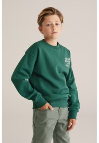 Zelena majica s okruglim ovratnikom, s prednjim logom "BLUE RIDGE" u bijeloj boji. Uparena s lakim zelenim hlačama, opuštenog kroja, s rebrastim manžetama.