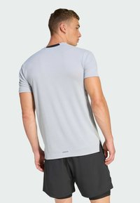 Camiseta deportiva gris claro con mangas cortas, fabricada con un tejido suave y con un dobladillo lateral dividido; combinada con pantalones cortos negros. Logotipo "AEROREADY" en la parte inferior.
