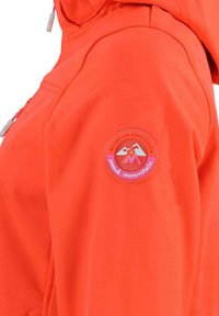 Veste softshell orange avec un patch circulaire portant le logo "Peak Mountain" et un graphisme de montagne sur la manche gauche.