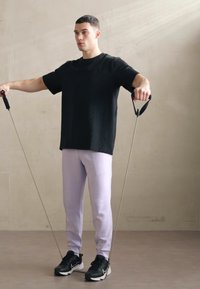 Black short-sleeve shirt, light purple sweatpants, and black athletic shoes. He holds exercise bands in both hands. Neutral background.