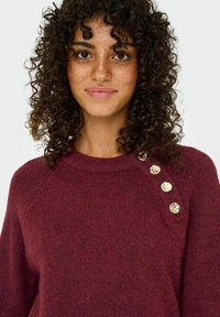 Pull maroon en maille avec un col rond côtelé, comportant quatre boutons métalliques dorés sur l'épaule gauche. Tissu texturé avec une finition douce.