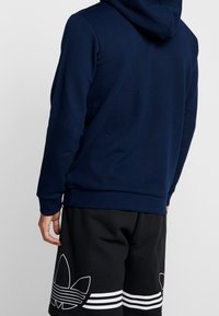 Man i en marinblå hoodie och svarta shorts med stor vit Adidas-logotyp och ränder, sedd bakifrån mot en enkel bakgrund.
