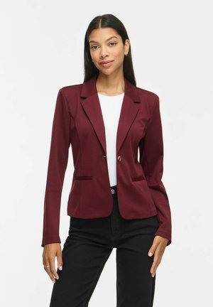 Kvinde med langt mørkt hår iført en tætsiddende burgundy blazer over en hvid top og sorte bukser, stående mod en ensartet lys baggrund.