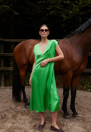 Kvinde i knaldgrøn kjole og solbriller står med den ene hånd i siden ved siden af en brun hest på et udendørs sandet område.