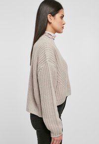 Ljusbeige stickad tröja med hög hals, sänkta axlar och en boxig passform. Har texturerade mönster och ribbade detaljer.