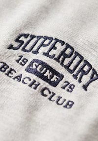 Textil gris con texto bordado en azul marino que dice "SUPERDRY 19 SURF BEACH CLUB". El bordado es elevado y la tela parece suave.
