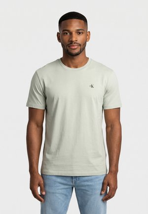 Giovane uomo che indossa una maglietta Calvin Klein beige chiaro e jeans azzurri, in piedi su sfondo bianco uniforme.