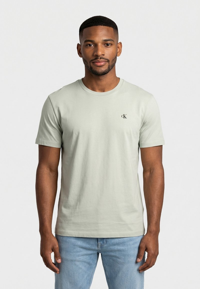 Jeune homme portant un t-shirt Calvin Klein beige clair et un jean bleu clair, debout devant un fond blanc uni.