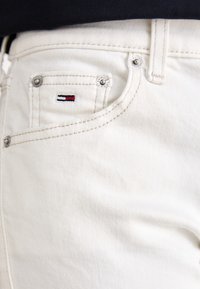 Vita denimjeans med en framficka som har en liten broderad logotyp i röd, vit och blå. Synlig metallprogramvara och sömnadsdetaljer.