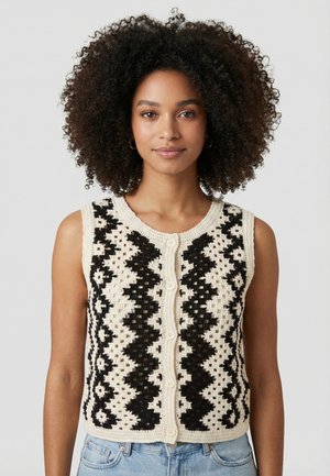 Femme aux cheveux bouclés naturels portant un gilet en crochet crème sans manches avec des motifs géométriques noirs et un jean bleu clair, sur un fond uni.