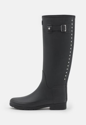 Bottes de pluie noires en caoutchouc montant jusqu'aux genoux, avec une boucle latérale et des clous argentés le long de la couture, dotées d'une semelle texturée pour une meilleure adhérence.