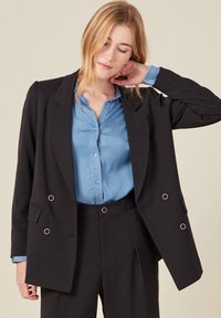 Blazer noir à double boutonnage avec des accents dorés, porté sur une chemise à boutons bleu clair. Présente des revers crantés et une coupe décontractée.