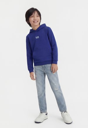Smiling boy wearing a blue EA7 hoodie, light ripped jeans, and white sneakers, standing against a plain white background.