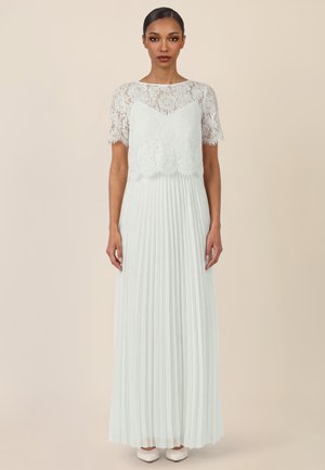 Frau steht mit dem Gesicht nach vorne, trägt eine weiße Spitzenbluse und einen langen plissierten weißen Rock mit weißen spitzen Schuhen.