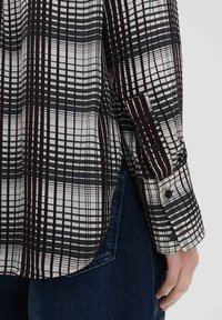 Camicia a scacchi bianca e nera, con maniche lunghe decorate da bottoni e un orlo ventilato, realizzata in tessuto leggero.