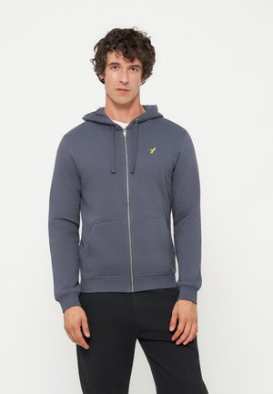 Jeune homme aux cheveux bouclés portant un sweat à capuche bleu zippé avec un logo jaune et un pantalon noir, debout devant un fond uni.