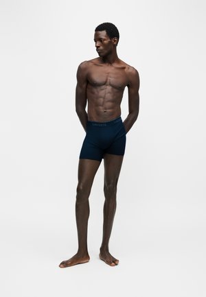Homme musclé debout pieds nus portant un boxer Lacoste bleu foncé, regardant vers sa gauche sur un fond clair uni.